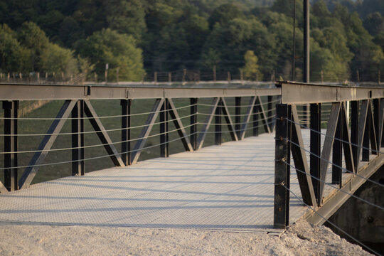 A sturdy black metal bridge with diagonal supports spans over calm water, leading toward a backdrop of green forest. The bridge&rsquo;s geometric design contrasts with the organic shapes