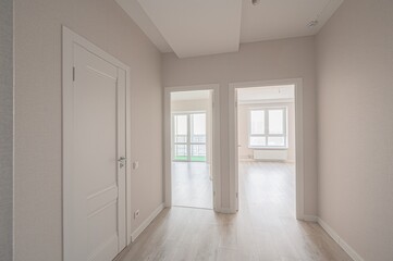 bright hallway with two open doorways leads into sunlit rooms. A closed white door is on the left. Walls are beige, floor is light wood