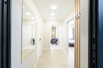 bright hallway with light beige walls, a large mirror, and light-colored tiles. An open doorway...