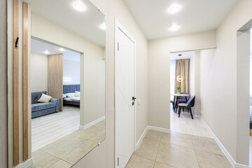 Modern apartment interior featuring a hallway with beige walls and tile flooring. Open doorways lead to a bedroom and dining area. A large mirror reflects the bedroom space