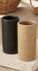 Two cylindrical, speckled, light-tan and dark-grey/black, decorative vessels sit on a light-gray stone surface