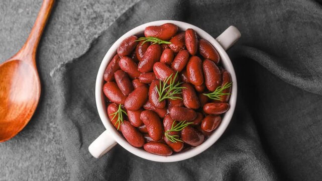 Freshly cooked red kidney beans fill a white bowl, presented beautifully with green herbs on a dark table, next to a wooden spoon. 