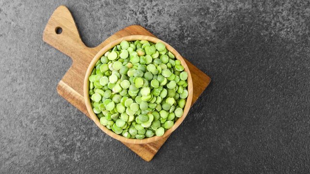 Overhead view of green split peas filling a natural wooden bowl on a small cutting board against a dark textured background. 