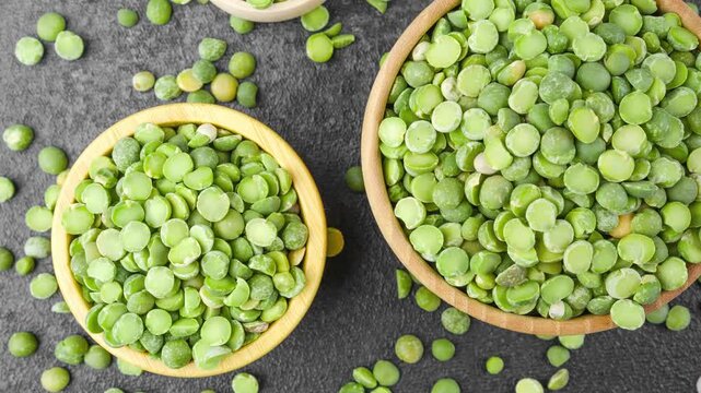 Bright green dried peas fill wooden bowls and scatter on a dark surface, creating a vibrant display of healthy plant based food ingredients. 