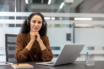 Fototapeta premium Professional woman sitting at desk, wearing headphones, working on a laptop in a modern office setting. The atmosphere reflects a productive environment, expressing focus, confidence, and dedication.