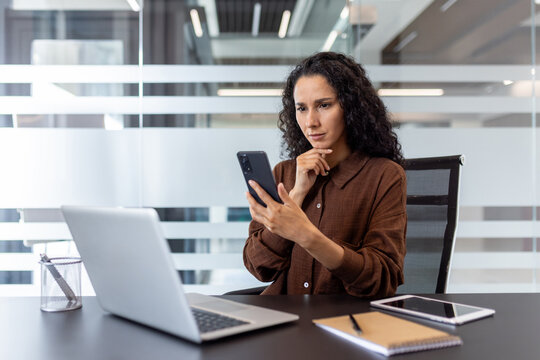 Businesswoman using a smartphone while working on a laptop in an office setting, surrounded by modern design and professional tools, reflecting focus, productivity, and digital communication