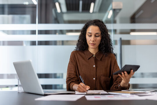 Confident businesswoman sitting at a desk, using a laptop and calculator, analyzing business data in a contemporary office. She appears diligent and focused on her work, portraying productivity and