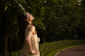 Beauty happy face woman looking up on sunset nature colors standing under the tree in a city street park at white column. Female brunette model relax in summer day with smiling face