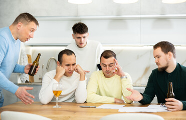 Upset worried man sitting at table in home kitchen with papers in hands, reading unexpected medical results with group of male friends solacing and comforting him ....