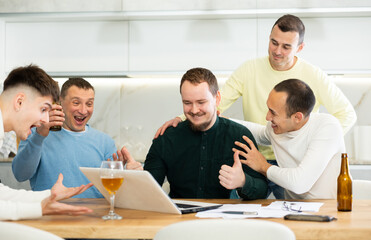 Group of men watching sports match online on laptop during friendly meeting with beer at home, excitedly celebrating winning score in game ..