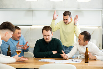 Upset worried man sitting at table in home kitchen with papers in hands, reading unexpected medical results with group of male friends solacing and comforting him ....