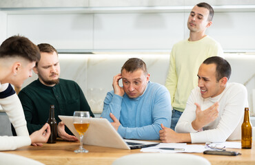 Group of men gathered for friendly meeting with beer at cozy home kitchen, trying to work through challenging problem, viewing laptop screen with concerned expressions and gestures ..