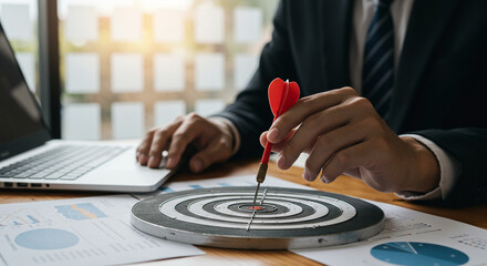 Businessman Achieving Goals:  Close-up Shot of Hands Placing Dart in Bullseye on Dartboard, Laptop and Financial Charts Present.
