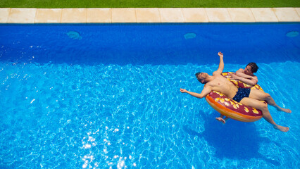 Young couple relaxing on a pastry ring pool float in clear blue water on a sunny day, the girl watches as the boy rests with his arms open with his head partially in the water