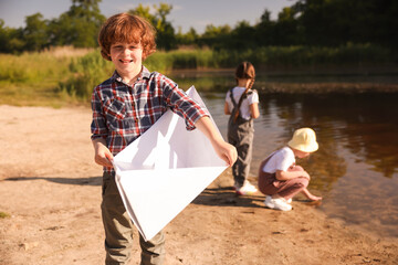 Children in nature, selective focus. Cute little boy with paper boat by lake