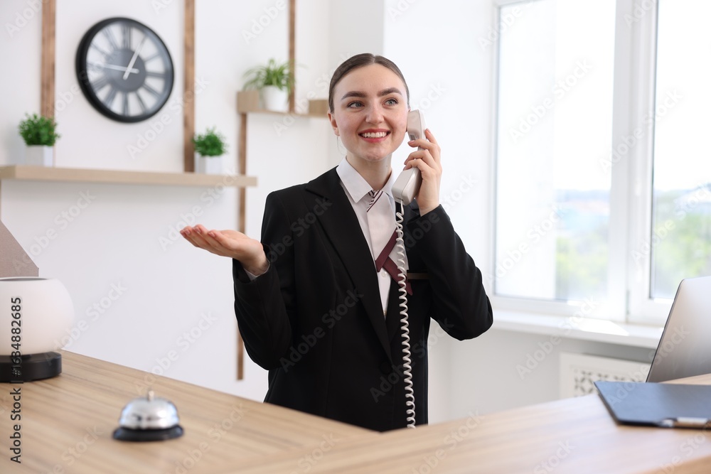 Poster smiling receptionist talking with client by phone at reception desk in hotel