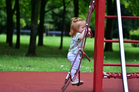 Red-haired toddler girl climbing a red rope ladder on a playground. Determined expression shows early childhood exploration, physical development, and outdoor play in a green forest park.