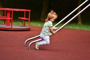Red-haired toddler sliding down a metal slide while holding on with both hands. The child is...