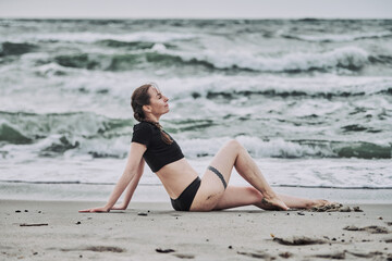 Young woman with brown hair practices yoga on baltic sea beach. Overcast sky, gentle waves create calming backdrop. Reflective, serene atmosphere