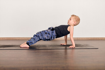 Child with blonde hair performs yoga on mat, wearing casual outfit. Indoor studio with wooden floor...
