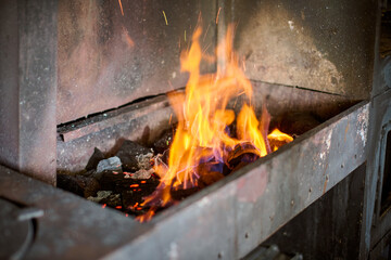 Intense flames rise from forge, casting warm glow in metal workshop. Soot-covered walls and scattered coal create authentic industrial atmosphere. Vibrant orange tones dominate scene