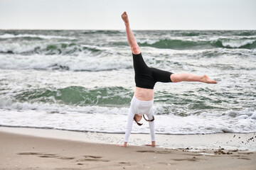 Young female with red hair executes cartwheel on sandy baltic shore. Overcast skies and gentle...