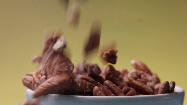 Sweet and tasty pecans and savory hickory native pecan nuts fall in slow motion slo mo into a white bowl full of snacks, crumbs, and flavor on a colorful studio lighting background