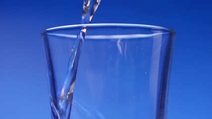Glass of water on a deep blue background in colorful good lighting studio as liquid in super slo mo slow motion drips and pours into drinking container and bubbles up to the top of the transparent cup - Powered by Adobe