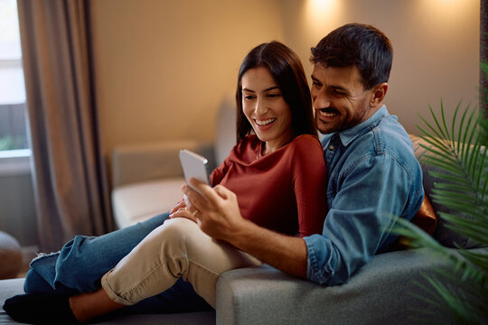 Happy couple using cell phone while relaxing on sofa at home.