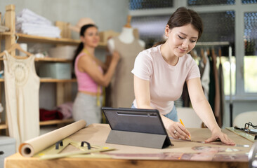 In workshop, girl designer draws diagram of future clothing product on paper and refers to image of...