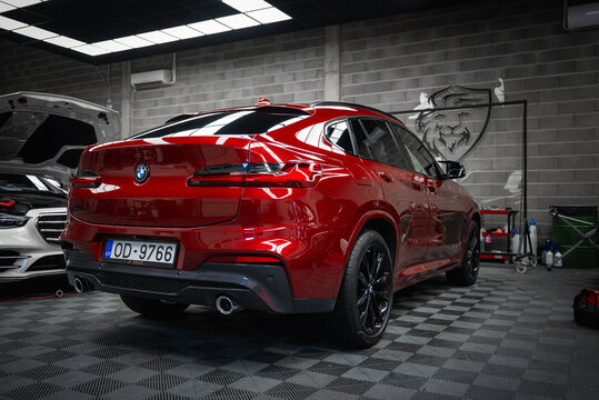 A red BMW X4 with a glossy finish is parked in a modern garage with tiled floors. A white Mercedes G Class with its hood open is partially visible nearby.