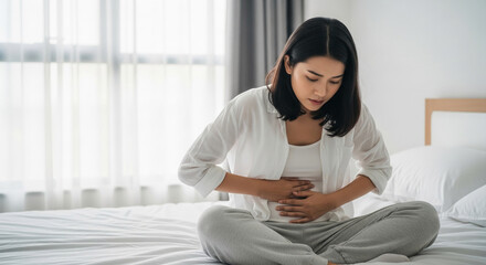 Woman in white shirt and gray pants sits on bed, hands holding stomach, expressing discomfort or pain. Image depicts unwellness, possibly stomach ache or menstrual cramps