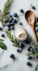 Overhead shot of a small jar of creamy substance surrounded by blueberries, rosemary, lavender, and sea salt on a marble surface, next to a wooden spoon