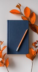 A navy blue notebook rests on a white background, adorned with vibrant orange autumn leaves and branches, a light-colored pencil resting atop