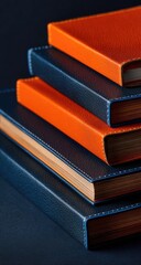 A close-up of four hardcover books stacked; two are dark blue, two are burnt orange;  they have visible gilded edges and subtly textured covers against a dark background