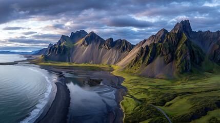 Jagged peaks rise above verdant slopes meeting a dark sand beach and calm ocean under dramatic clouds