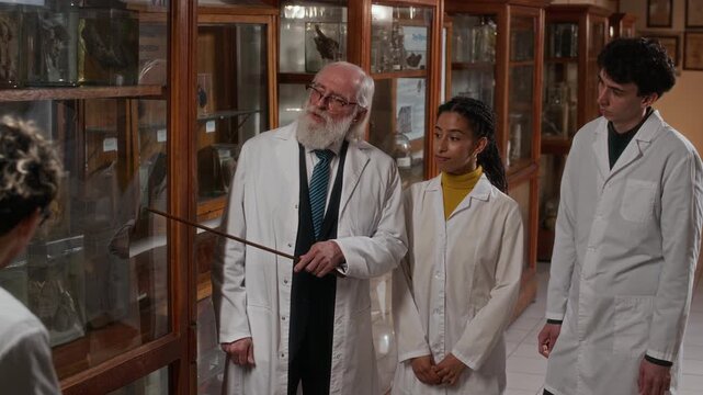 Mature Caucasian male professor in white coat guiding museum tour for motivated multiethnic medical interns in scrubs listening to him attentively