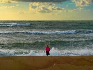 A person in a bright pink jacket stands at the edge of a windy seashore, facing the powerful waves of a stormy sea. The dramatic clouds and golden hour sky create a striking backdrop