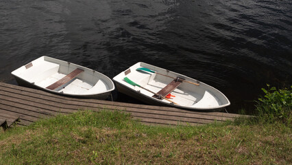 Two small white rowboats tied to a wooden dock at the edge of a calm, dark river. One boat is equipped with colorful paddles—green and turquoise—and bright orange life jackets.
