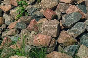 A pile of large, rough granite stones in various shades of red, gray, and brown, surrounded by green grass and small plants.