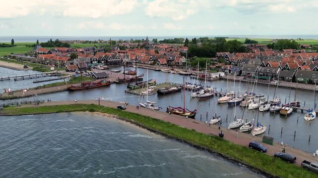 Aerial view of a charming Dutch harbor town with traditional red-roofed houses, moored sailboats, and calm waters surrounded by green fields and open sky, creating a serene coastal scene.