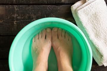 Women's feet in a basin of water on a dark wooden floor and a towel, home foot washing procedure,...