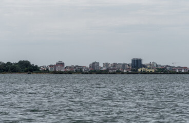 Naklejka premium Grado, Italy - July 24, 2025: Panoramic view of Grado from Barbana island, with modern buildings rising above the lagoon on a cloudy day over calm northern waters.
