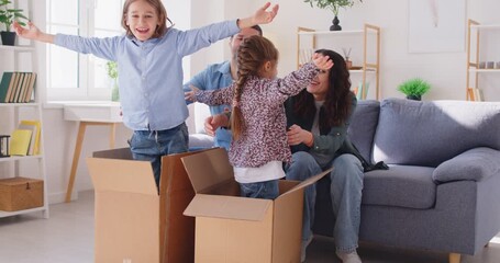 Cheerful parents hugging their two laughing children appearing from moving boxes. Happy smiling family having fun and embracing while unpacking moving boxes, celebrating buying or renting new home. - Powered by Adobe