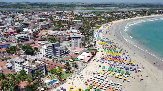 Aerial view of Pontal do Cupe Beach in Porto de Galinhas, Ipojuca, Pernambuco, Brazil. A beautiful coastline with turquoise water, colorful umbrellas, and a vibrant coastal town under a cloudy sky.