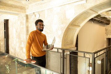 Confident black professional holding laptop and notebook while standing in bright office corridor