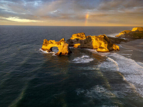 Aerial view of the rugged, golden cliffs of Wharariki Beach meet the turbulent, deep blue sea under a pastel sky kissed by a faint rainbow, P&Aring;&laquo;ponga, Tasman Region, New Zealand.