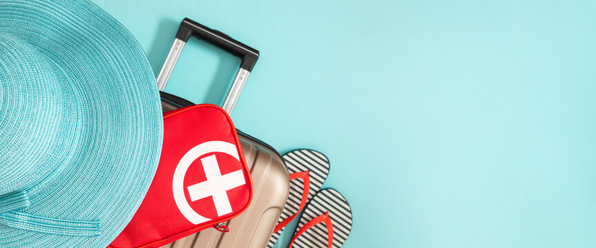 Summer travel concept: suitcase, flip-flops, hat and red first aid kit on blue background. Banner.