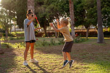 Obraz premium Happy blond-haired boy joyfully jumping to pop soap bubbles in a sunny park, while a smiling woman in casual clothes creates bubbles with a wand, surrounded by trees and warm afternoon light