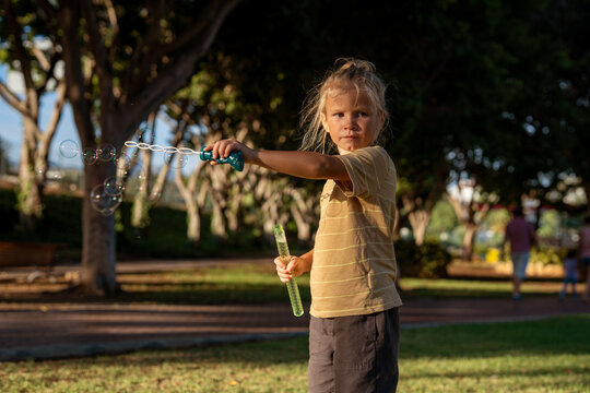 Young blond boy with a focused expression blowing soap bubbles in a sunny park, holding a bubble wand in one hand and bubble liquid in the other, surrounded by trees and natural light